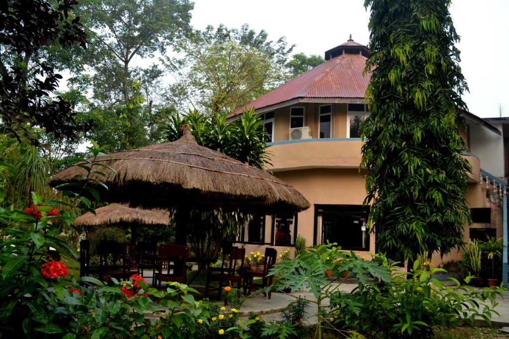 Distinctive round tower building with red metal roof and thatched-roof garden pavilions surrounded by flowering tropical gardens at Rhino Lodge & Hotel Sauraha Chitwan Nepal