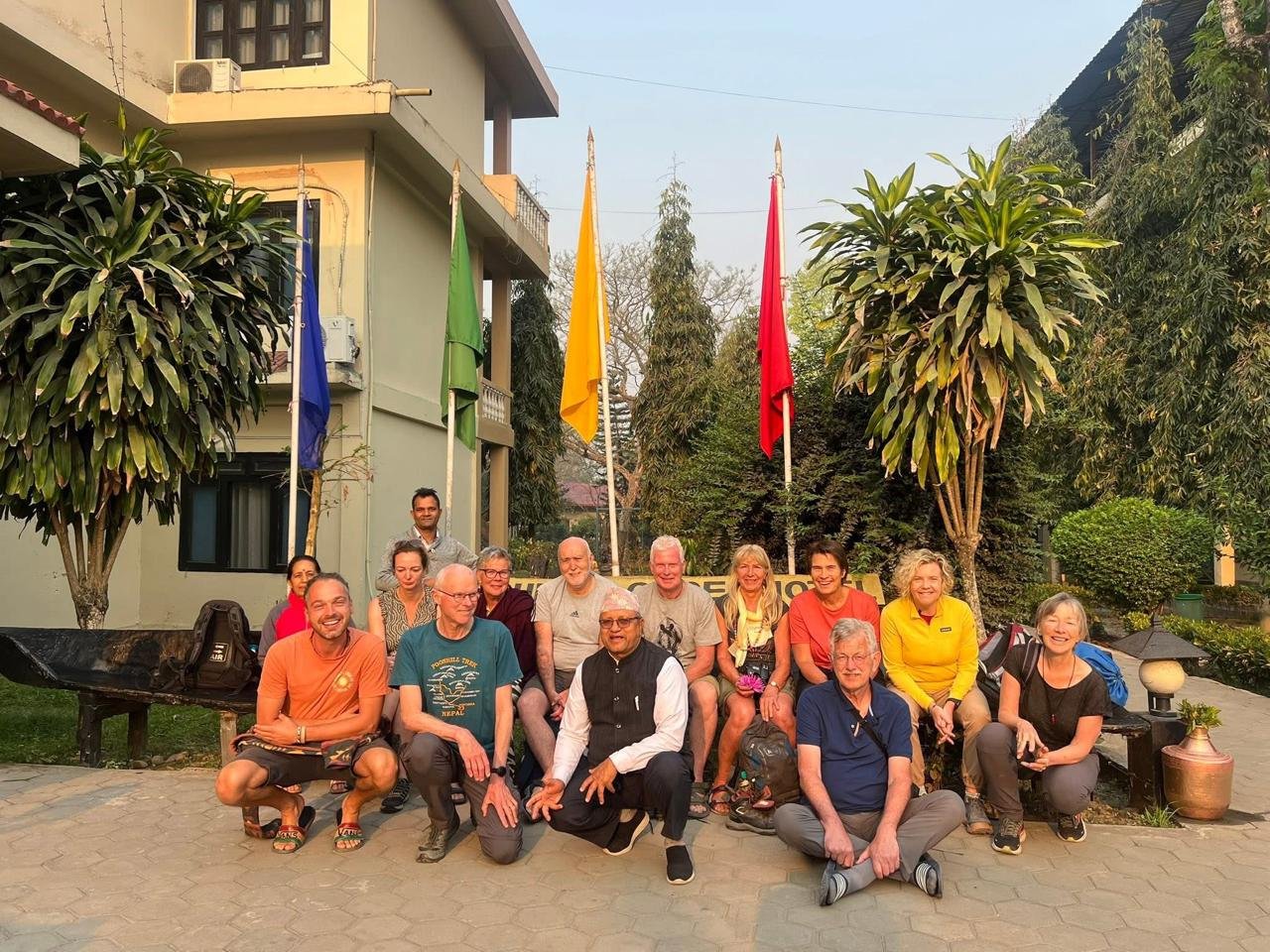 International tour group with local guide posing at Rhino Lodge & Hotel entrance in Sauraha Chitwan Nepal with colorful prayer flags and tropical landscaping