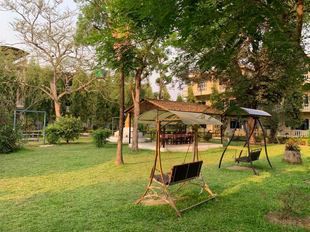 Lush garden at Rhino Lodge & Hotel Sauraha Chitwan featuring garden swings, covered gazebo dining area, and mature trees with hotel building backdrop