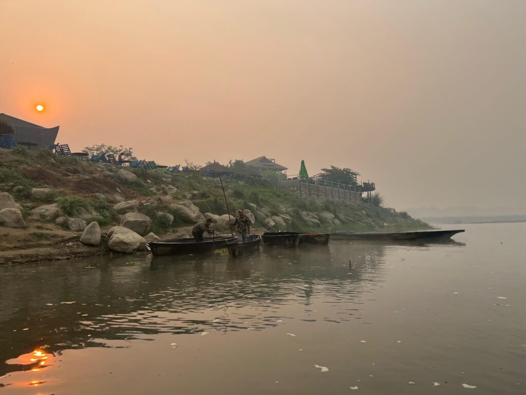 A calm riverside scene at sunset with small wooden boats, fishermen, and a rocky hillside lined with riverside lodges and seating areas.