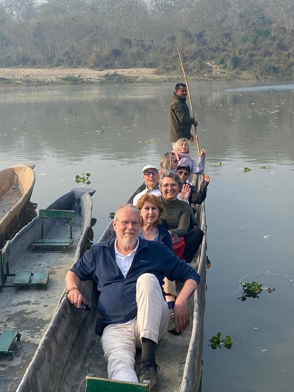 A group of tourists sits in a narrow wooden boat on a calm river, with a boatman standing at the rear holding a long pole.